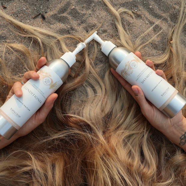 Two bottles of hair care product being used on wet hair on a sandy background
