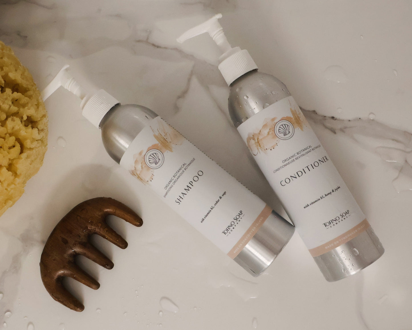 Two bottles of shampoo and conditioner with a scrubber and comb on a marble surface