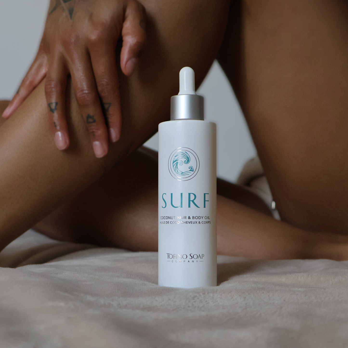 Skincare bottle labeled 'Surf' on a bed with a person sitting in the background.