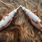 Two bottles of hair care product being used on wet hair on a sandy background