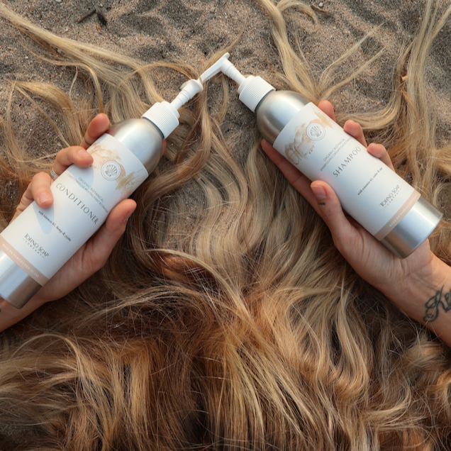Two bottles of hair care product being used on wet hair on a sandy background